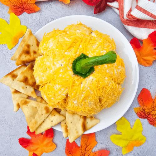 A pumpkin cheese ball with a green stem is served on a white plate with pita chips, surrounded by colorful artificial fall leaves and a striped towel.