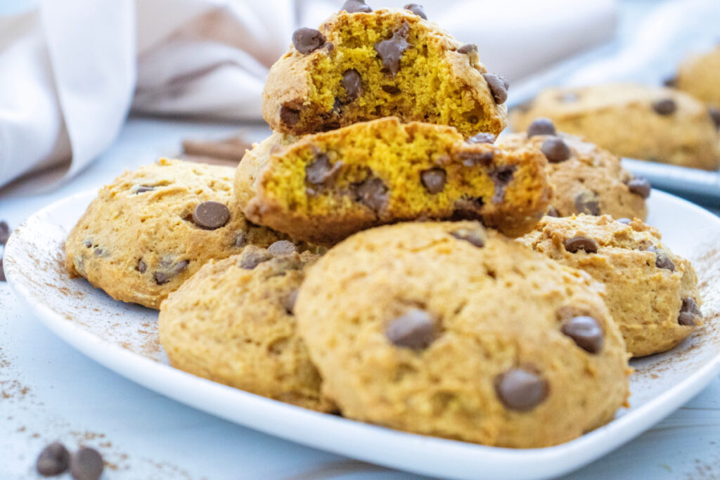 A plate of chocolate chip cookies, with one cookie split in half to show the soft, golden interior. Other whole cookies are scattered around the plate.