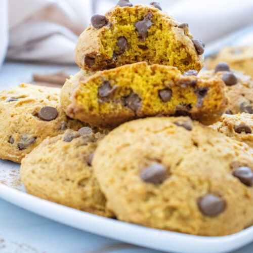 A plate of chocolate chip cookies, with one cookie split in half to show the soft, golden interior. Other whole cookies are scattered around the plate.