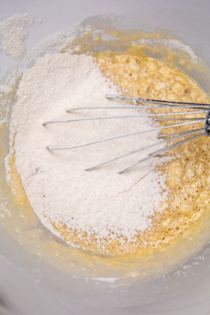 A metal whisk mixes flour into a bowl of wet ingredients, preparing a batter for pumpkin cinnamon rolls.