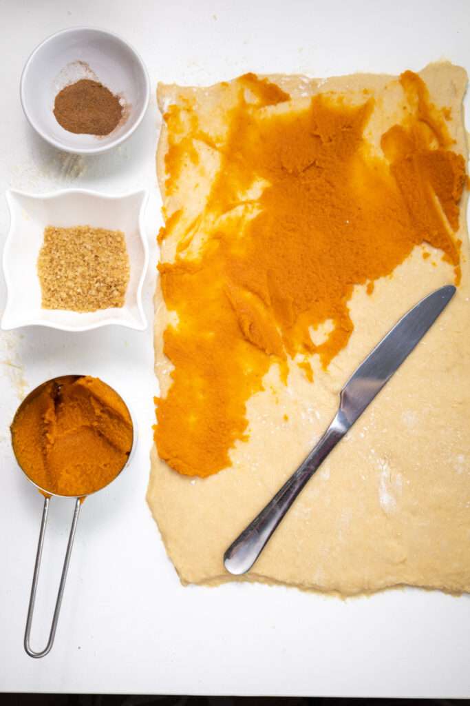 Rolled out dough for pumpkin cinnamon rolls is spread with pumpkin puree, next to a knife, a measuring cup with pumpkin, a bowl of spices, and a bowl of brown sugar on a white surface.
