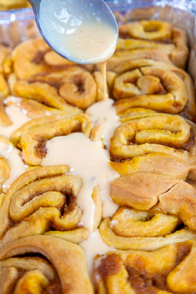 A spoon drizzles icing over a tray of freshly baked pumpkin cinnamon rolls.