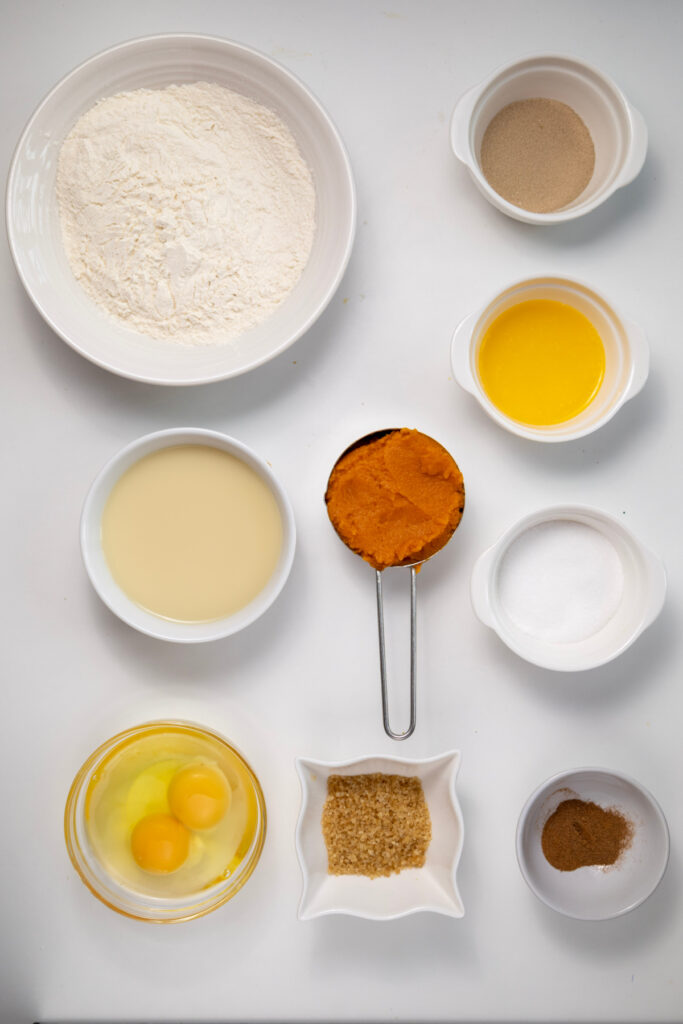 Top-down view of measured baking ingredients for pumpkin cinnamon rolls, including flour, yeast, melted butter, milk, pumpkin puree, sugar, eggs, brown sugar, and spices on a white surface.