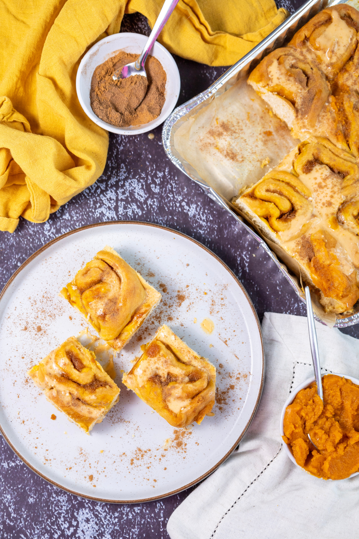 Three pumpkin cinnamon rolls are on a white plate, with more pumpkin cinnamon rolls in a baking dish. Nearby sit bowls of cinnamon sugar and pumpkin puree on a dark tabletop.