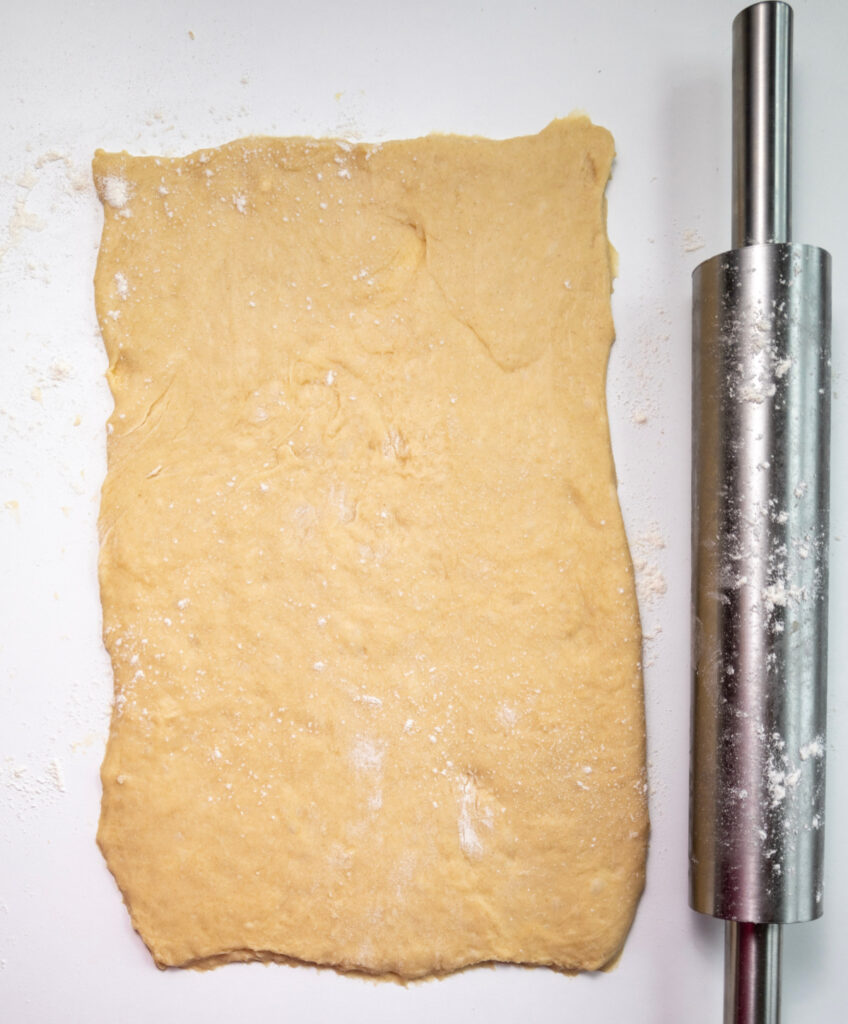 Rectangular sheet of raw dough rolled out on a white surface, ready to be transformed into pumpkin cinnamon rolls, with a metal rolling pin placed to the right.