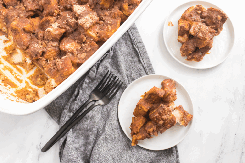A white baking dish with pumpkin french toast bake sits next to two small plates with servings, a grey cloth, and two black forks on a marble surface.