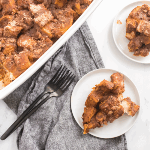 A white baking dish with pumpkin french toast bake sits next to two small plates with servings, a grey cloth, and two black forks on a marble surface.
