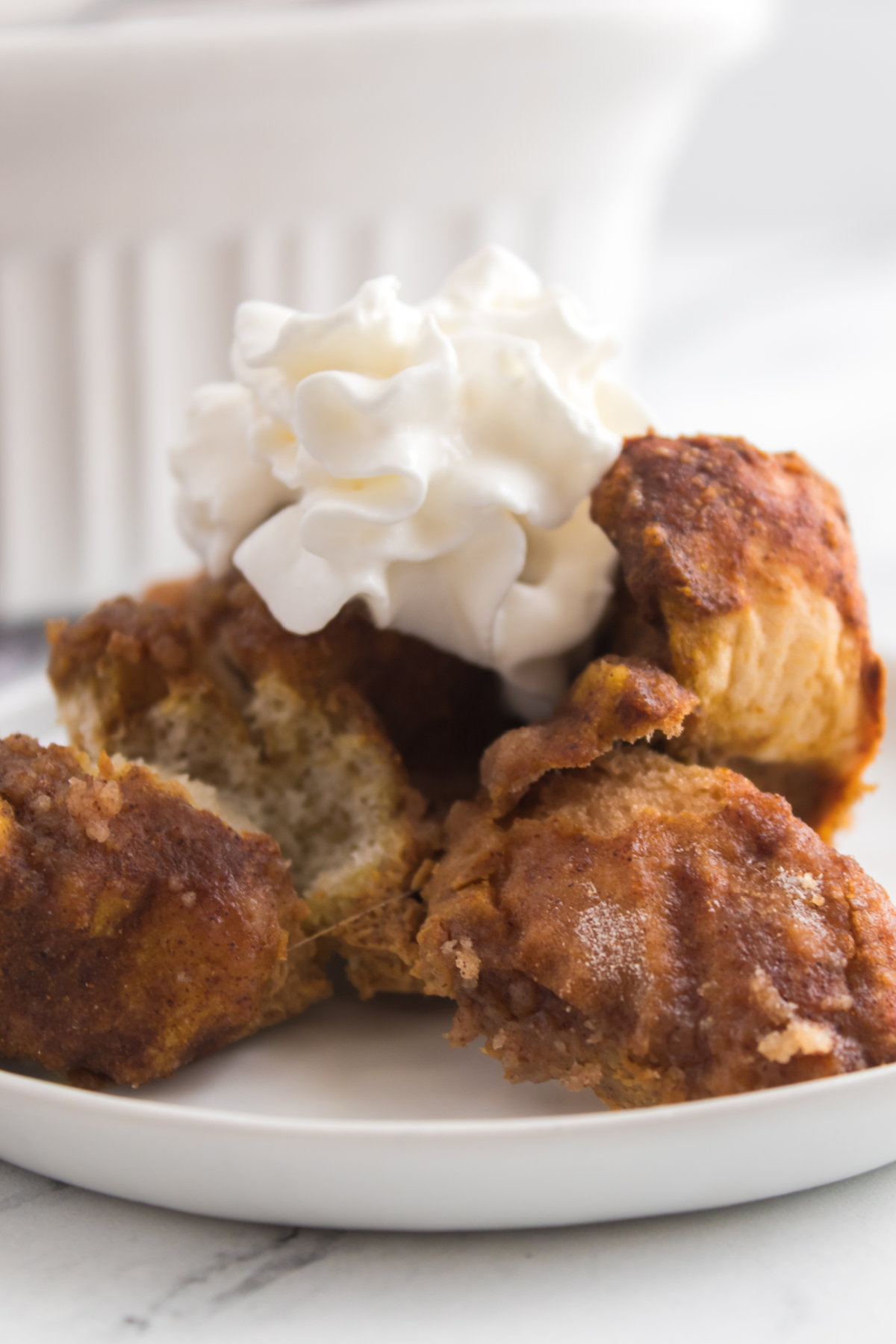 Four pieces of pumpkin French toast bake are stacked on a plate with a dollop of whipped cream on top; a white dish is in the background.