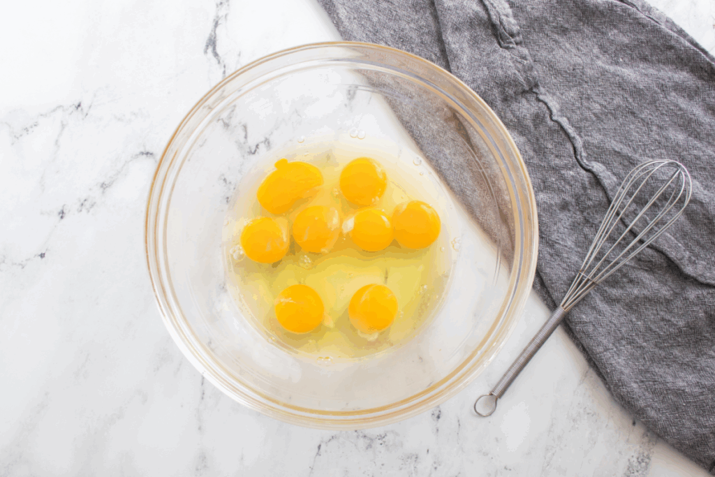 A glass bowl containing eight cracked eggs for a pumpkin french toast bake, with a metal whisk and a gray cloth nearby on a marble surface.