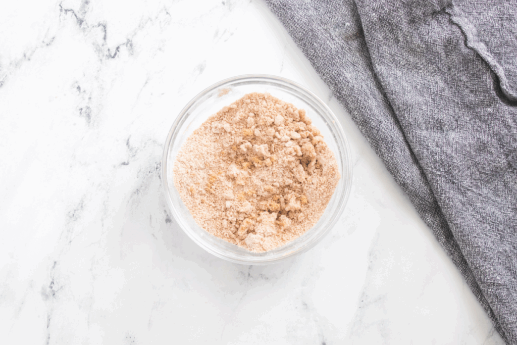 A clear glass bowl filled with a light brown, crumbly pumpkin French toast bake mixture sits on a white marble surface next to a gray cloth.