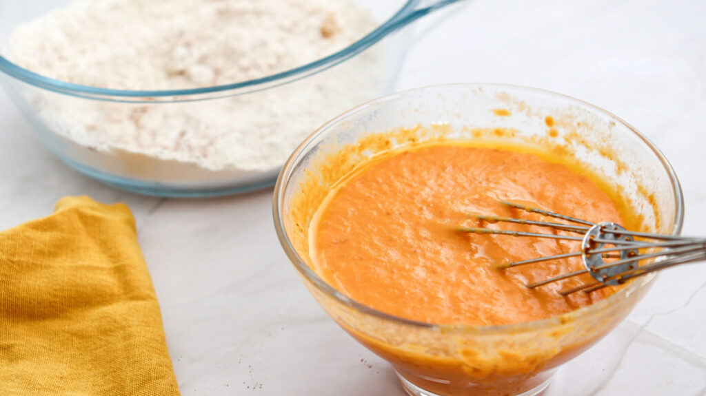 A glass bowl of orange batter with a whisk sits next to a bowl of flour mixture on a white surface with a yellow cloth nearby.