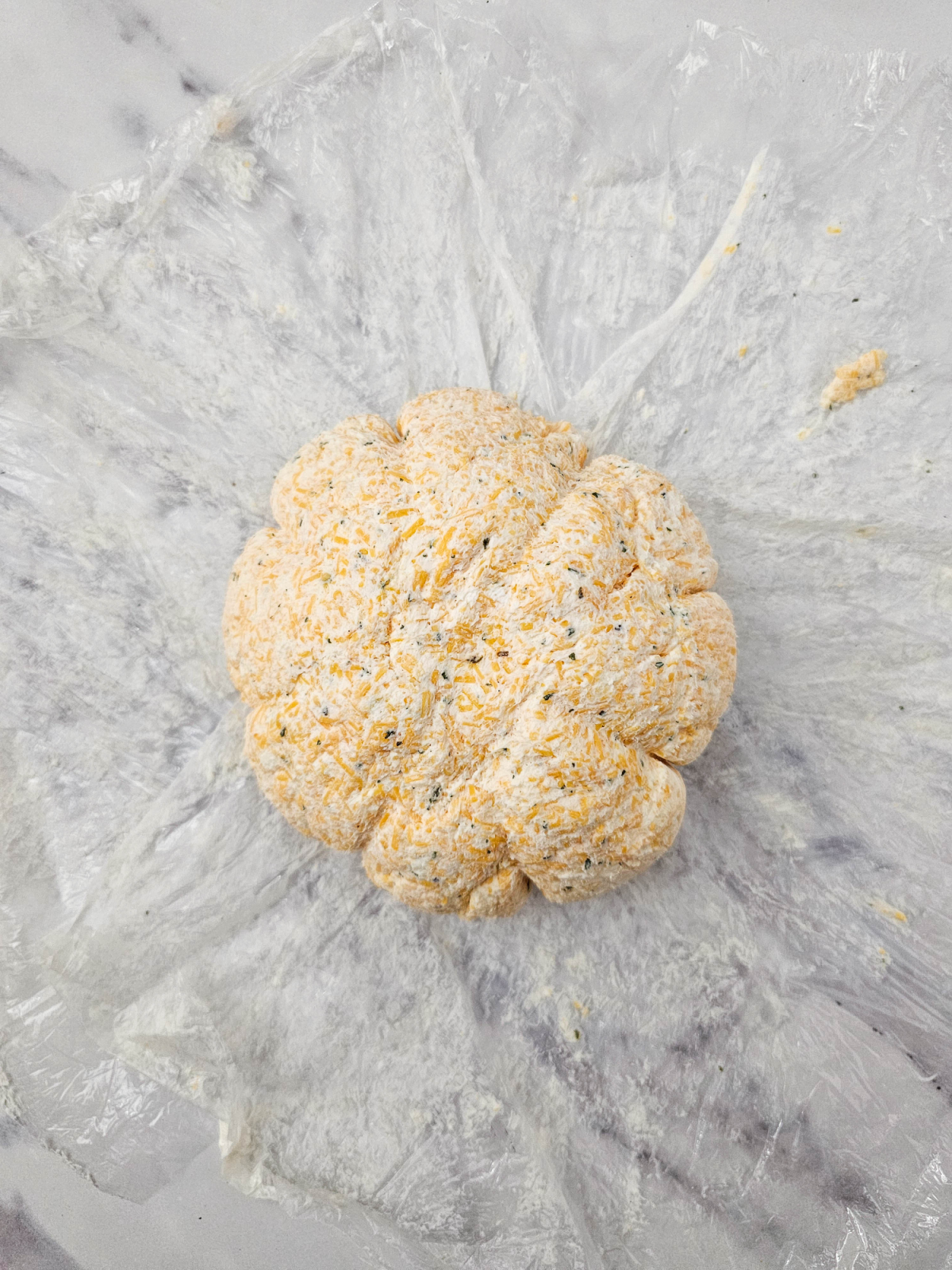 A round pumpkin cheese ball, blended with herbs and shaped into segments, sits on plastic wrap on a marble surface.