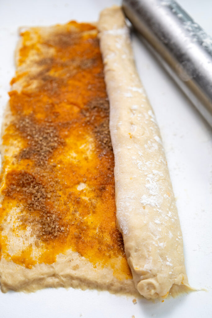 Dough for pumpkin cinnamon rolls is spread with cinnamon sugar and rolled up, with a rolling pin nearby on a white surface.
