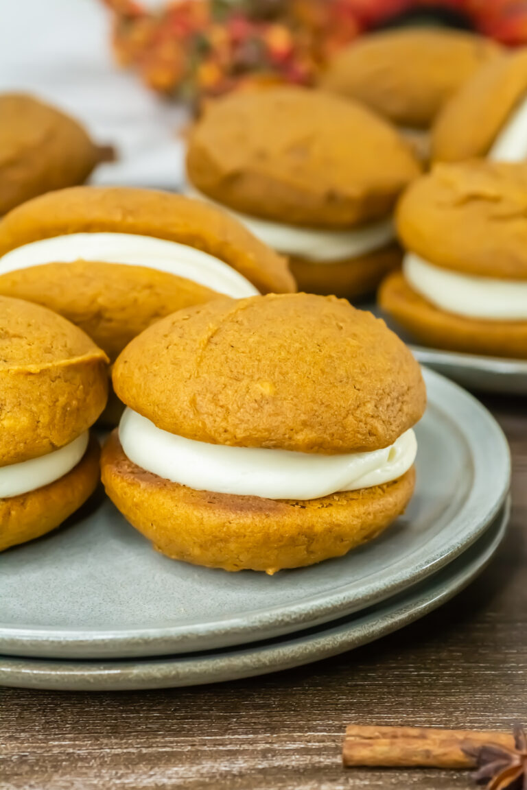 A plate of pumpkin whoopie pies filled with white cream, set on a wooden surface with more whoopie pies and fall decor in the background.