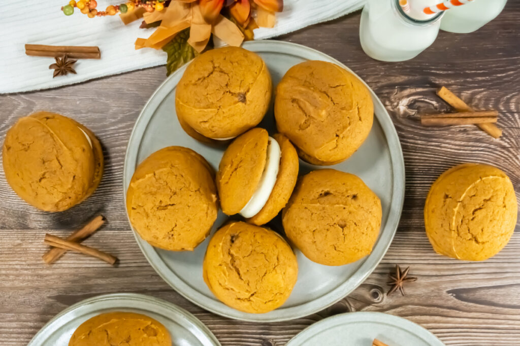 A plate of pumpkin whoopie pies, with one showing a cream filling, on a wooden table with cinnamon sticks and two bottles of milk.