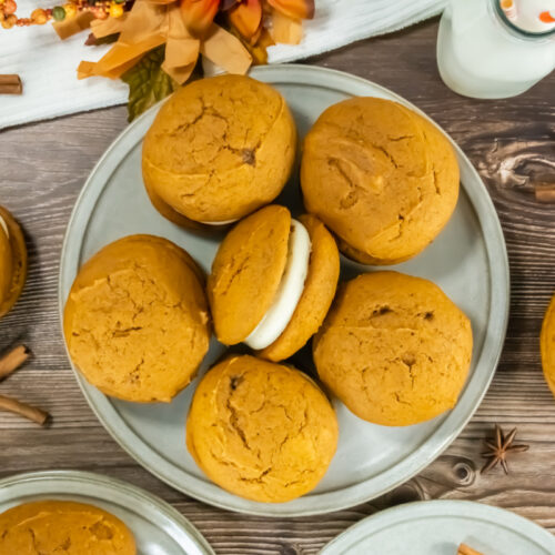 A plate of pumpkin whoopie pies, with one showing a cream filling, on a wooden table with cinnamon sticks and two bottles of milk.