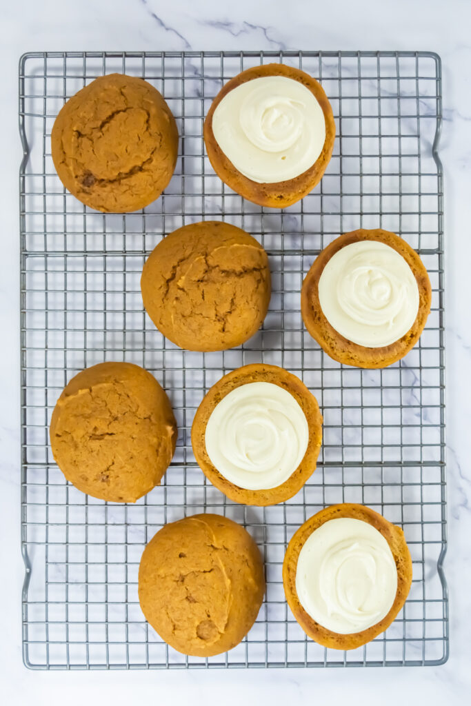 Eight pumpkin cookies, four with white icing and four without, arranged on a metal cooling rack on a marble surface.