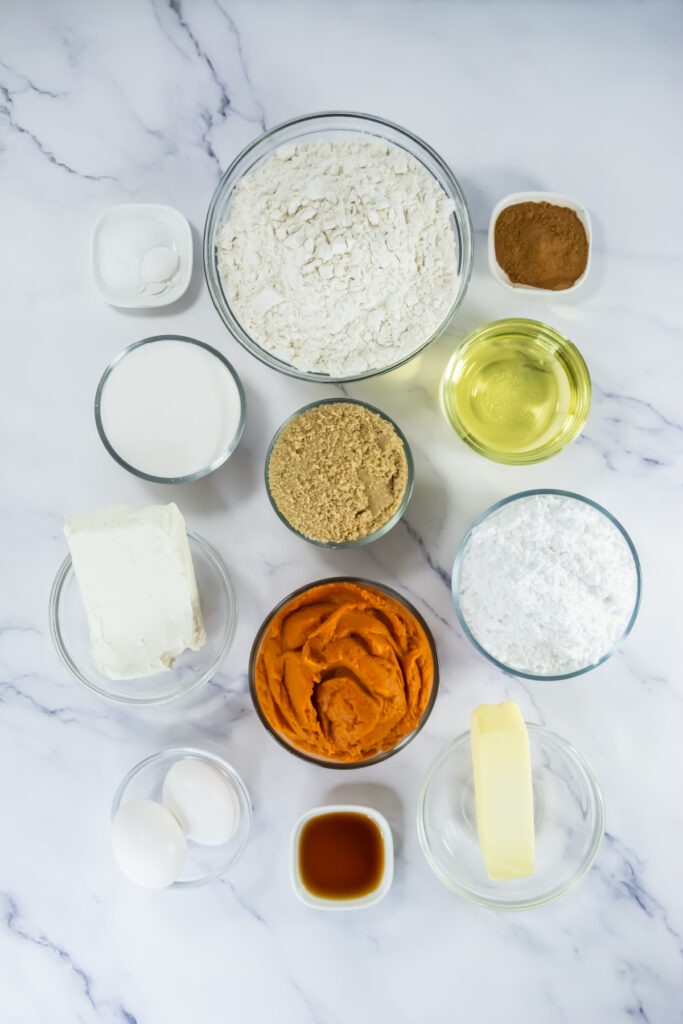 Assorted baking ingredients in glass bowls on a marble surface, including flour, sugar, brown sugar, pumpkin puree, cream cheese, eggs, butter, oil, cinnamon, vanilla, and powdered sugar.