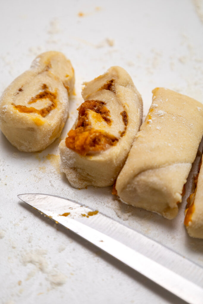 Unbaked pumpkin cinnamon rolls with a visible orange filling are sliced into pieces on a floured surface next to a metal knife.