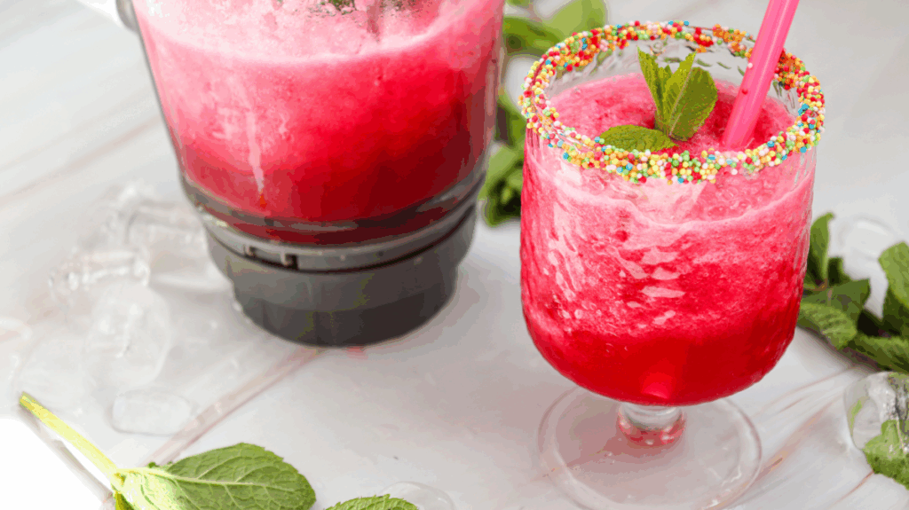 A vibrant watermelon slushie in a glass with a sprinkle-covered rim, mint garnish, and pink straw sits next to a blender of the same refreshing beverage on a white surface with ice and mint leaves.