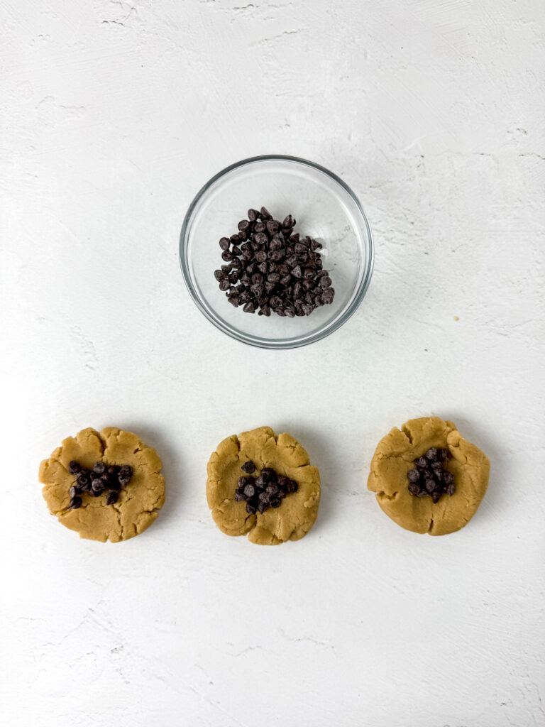 Three unbaked cookie dough balls with chocolate chips in the center are arranged in a row beneath a bowl of chocolate chips on a white surface.
