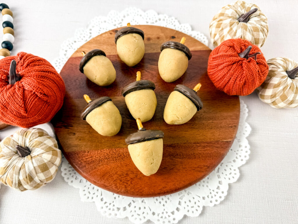 A wooden tray holds seven acorn-shaped cookies with chocolate tops and pretzel stems, surrounded by decorative fabric pumpkins on a white lace mat.