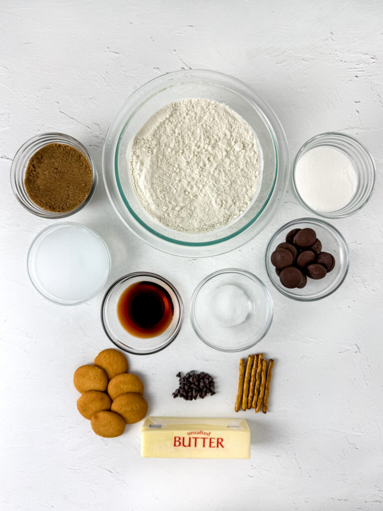 Top-down view of baking ingredients including flour, sugars, milk, chocolate, vanilla, salt, gingersnaps, chocolate chips, pretzel sticks, and a stick of butter arranged on a white surface.