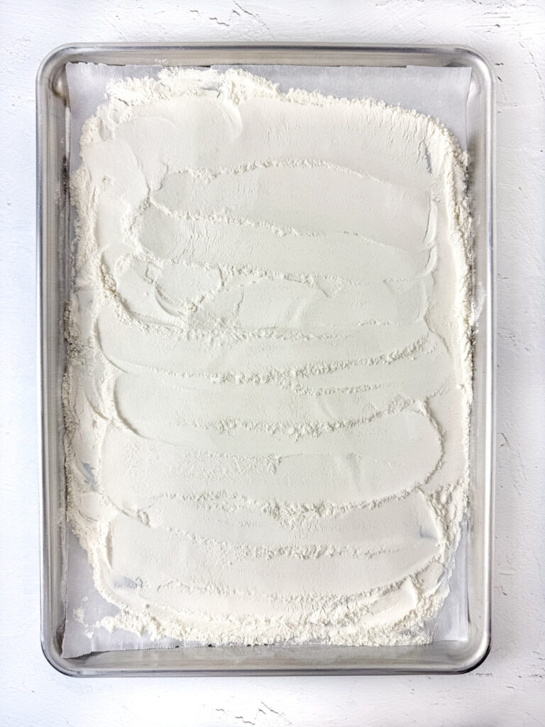 A metal baking sheet is evenly covered with a layer of white flour, spread out across the surface.
