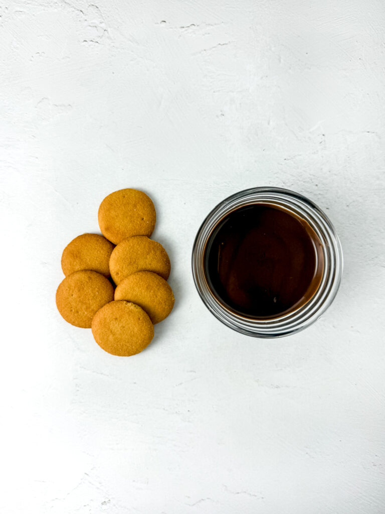 A glass of chocolate milk is placed on a white surface next to a small pile of round vanilla wafer cookies.
