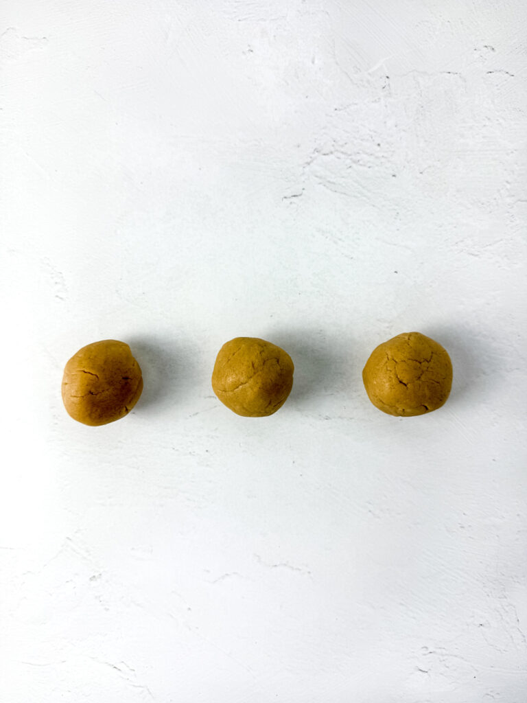 Three round, brown dough balls are arranged in a horizontal line on a white textured surface.