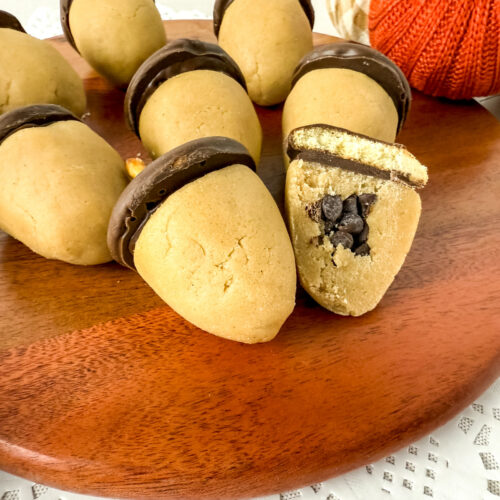 A wooden tray holds acorn-shaped cookies made of cookie dough and chocolate, with one cookie cut open to show chocolate chips inside. A knitted pumpkin decor is in the background.