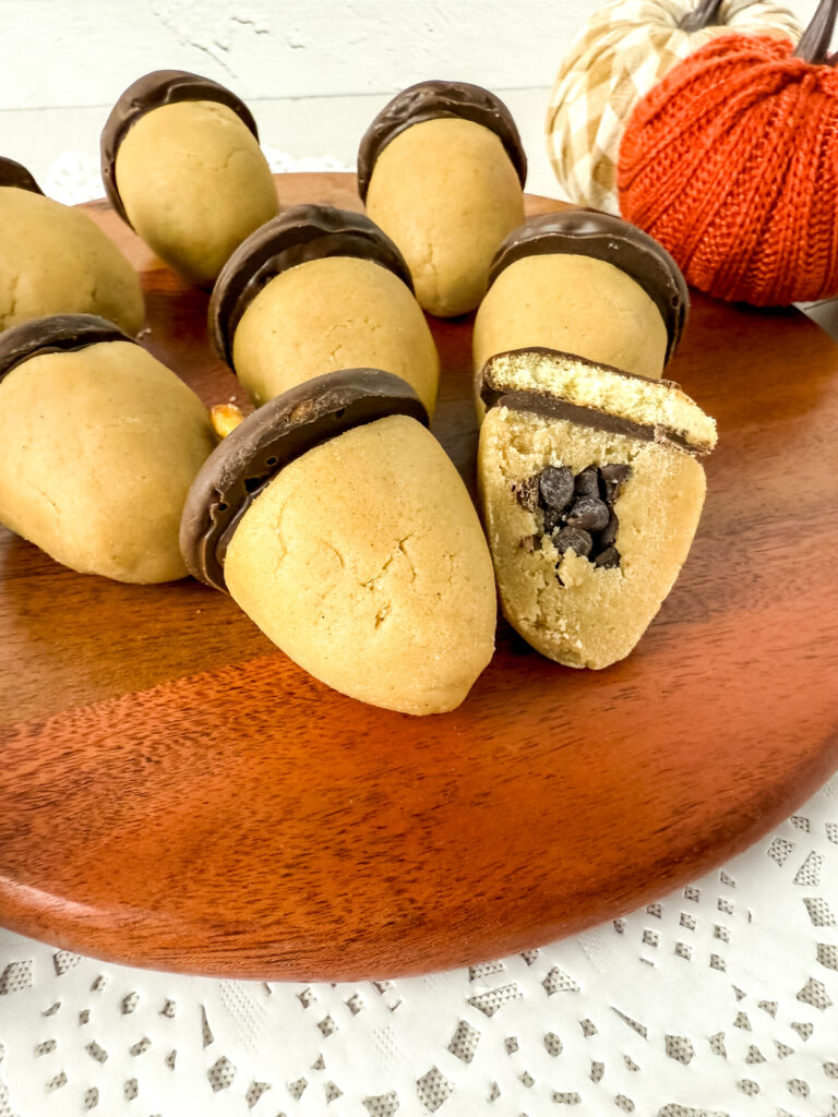 A wooden tray holds acorn-shaped cookies made of cookie dough and chocolate, with one cookie cut open to show chocolate chips inside. A knitted pumpkin decor is in the background.