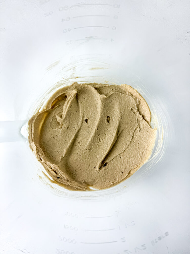 Thick, light brown batter is spread in swirling patterns inside a clear mixing bowl, viewed from above. Measurement markings are visible on the side of the bowl.