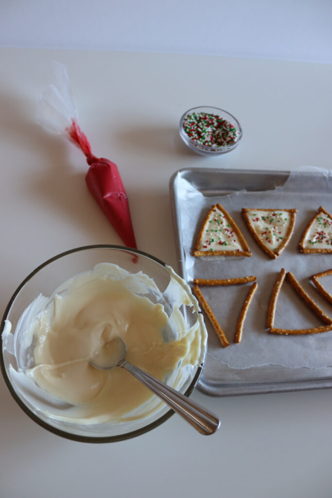 A bowl of melted white chocolate with a spoon, a piping bag, a small bowl of sprinkles, and a tray of decorated cookie wedges on parchment paper.