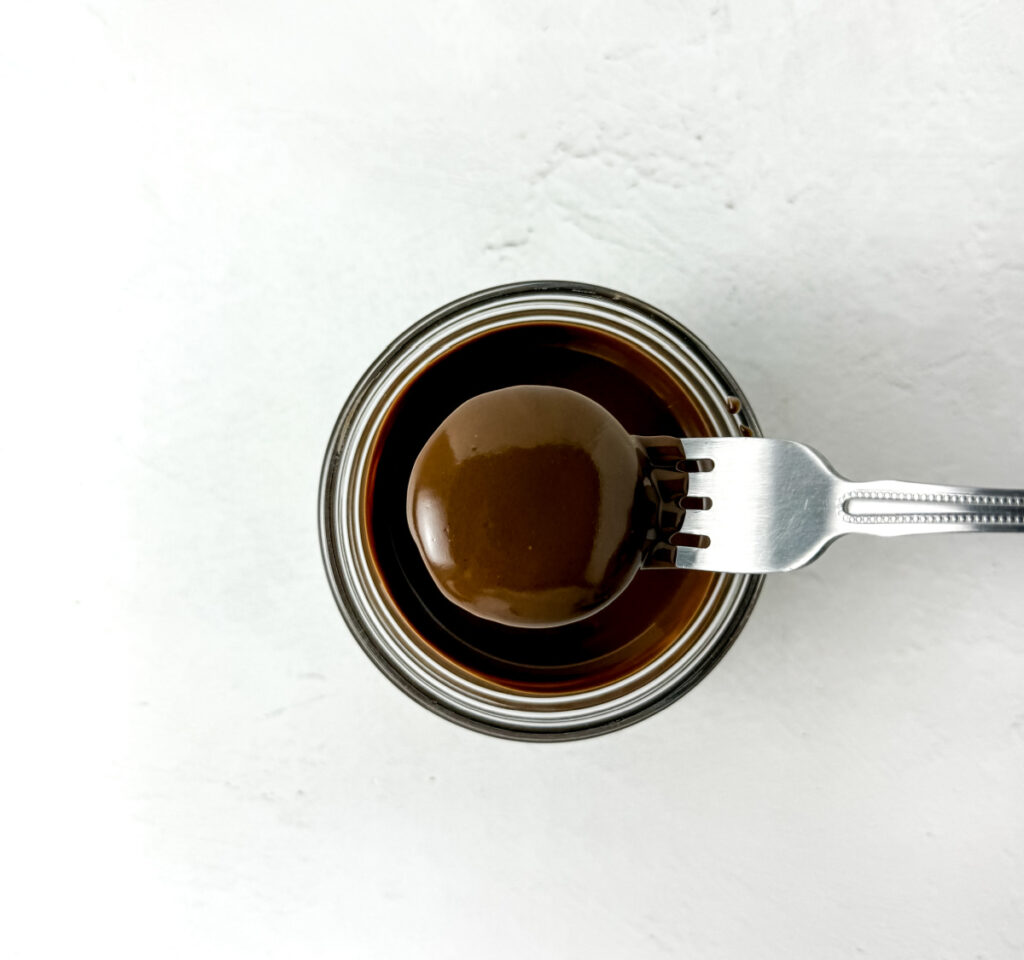 A fork holds a round chocolate-covered treat above a small glass jar filled with chocolate on a white background.