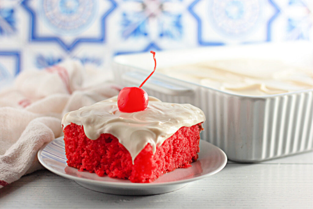 A slice of bright red cake with white frosting and a cherry on top sits on a plate, with the rest of the cake in a baking dish in the background.