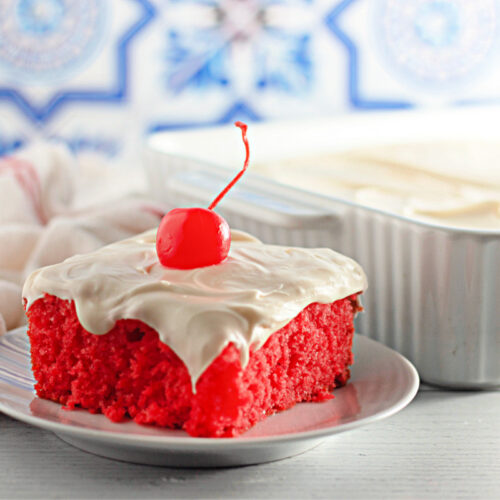 A slice of bright red cake with white frosting and a cherry on top sits on a plate, with the rest of the cake in a baking dish in the background.