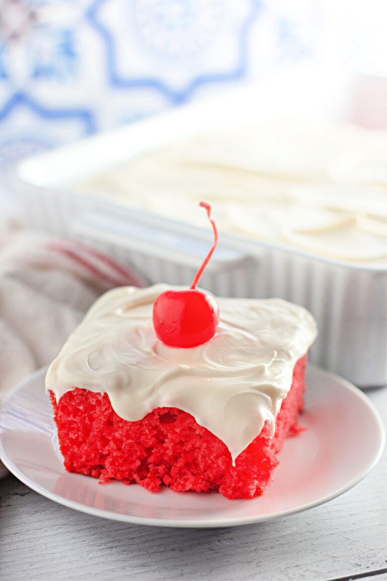 A slice of bright red cake with white frosting and a cherry on top sits on a white plate, with the remaining cake in a baking dish in the background.