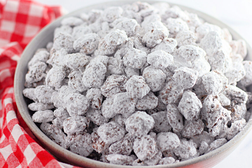 A bowl filled with chocolate-covered cereal pieces coated in powdered sugar, next to a red and white checkered cloth.