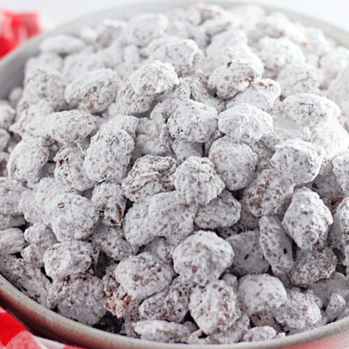 A bowl filled with chocolate-covered cereal pieces coated in powdered sugar, next to a red and white checkered cloth.
