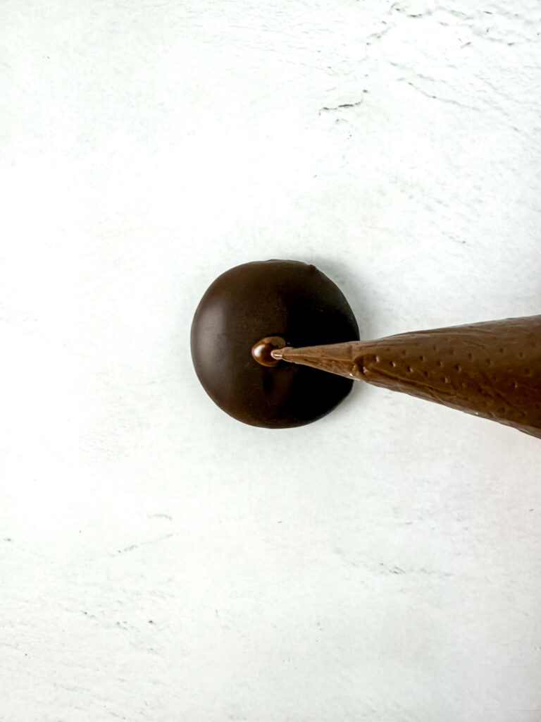 A piping bag decorates the top of a round, chocolate-covered treat on a white surface.