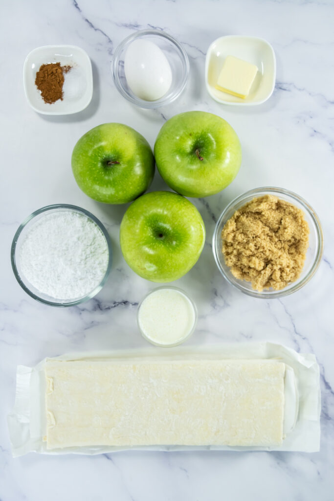 Ingredients for baking: three green apples, egg, butter, brown sugar, powdered sugar, milk, ground cinnamon, and sheet of puff pastry arranged on a white marble surface.