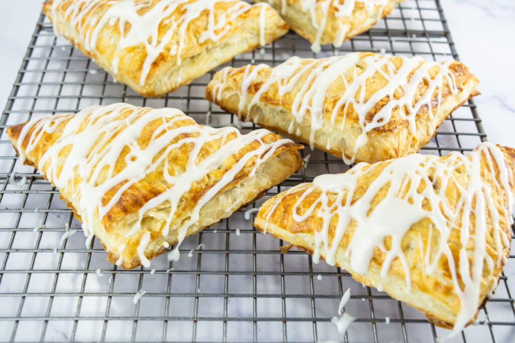 Four golden-brown pastry turnovers with white icing drizzle are cooling on a metal wire rack.