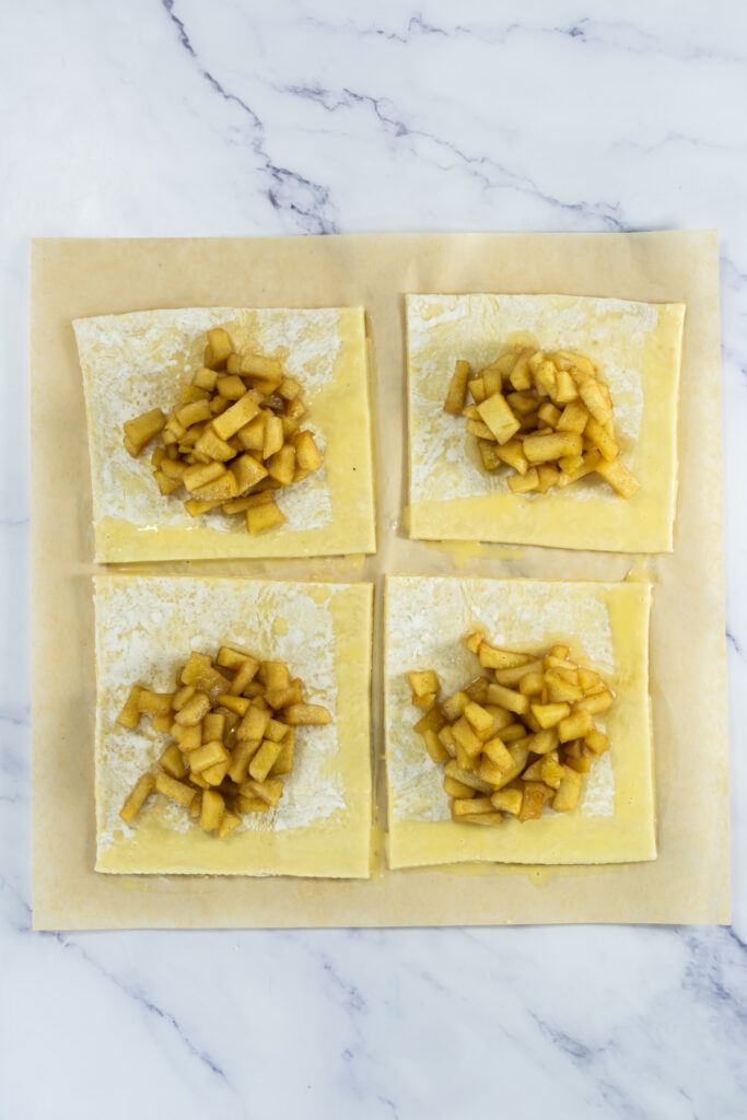 Four square pieces of pastry dough on parchment paper, each topped with a pile of diced apple filling, on a marble surface.