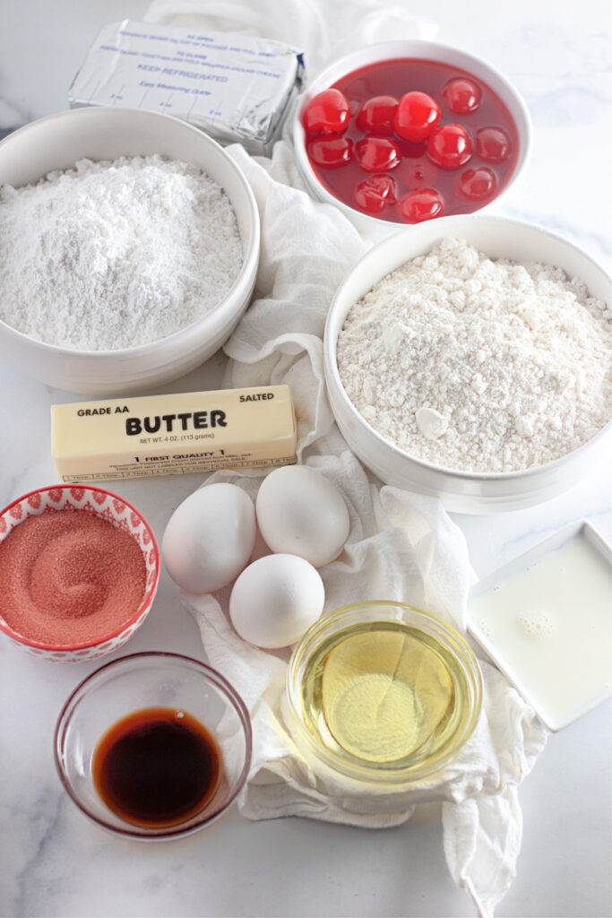 Ingredients for baking including flour, powdered sugar, cherries, butter, eggs, oil, vanilla, red sugar, and milk arranged on a white surface.