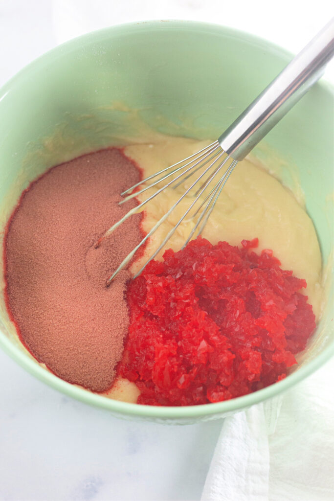 A mixing bowl with cake batter, red gelatin powder, and crushed red gelatin dessert, with a metal whisk partially submerged.