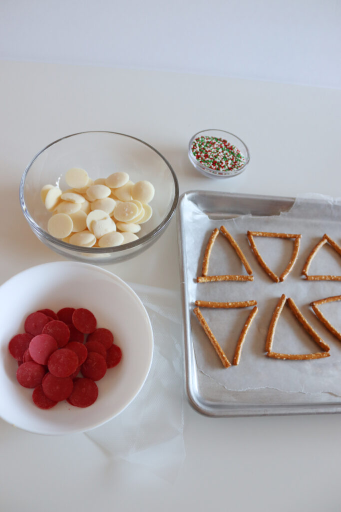 A bowl of white chocolate wafers, a bowl of red chocolate wafers, a small bowl of sprinkles, and a tray with pretzel triangles on parchment paper.