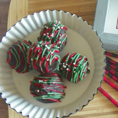 A white plate with four chocolate truffles decorated with green and red icing and sprinkles, placed on a wooden surface.