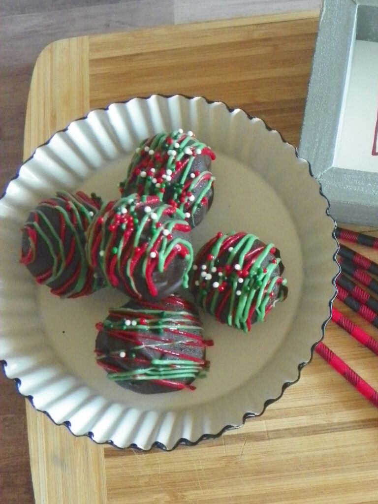 A white plate with four chocolate truffles decorated with green and red icing and sprinkles, placed on a wooden surface.