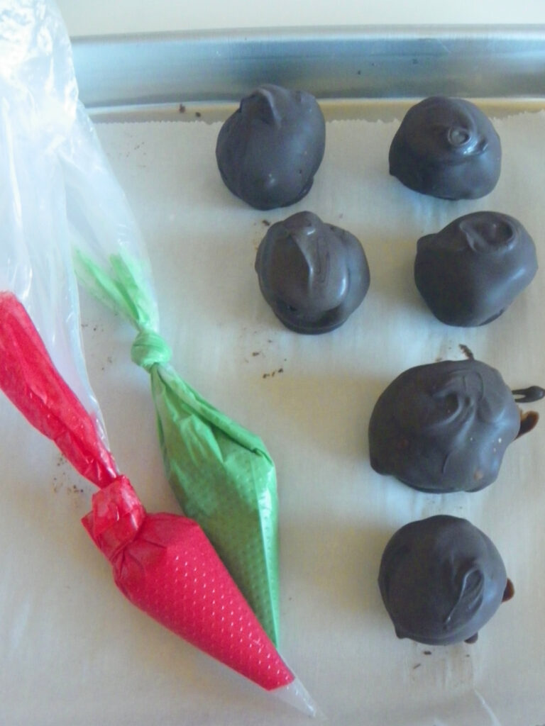 Six chocolate-coated treats on parchment paper next to two piping bags, one red and one green, prepared for decorating.